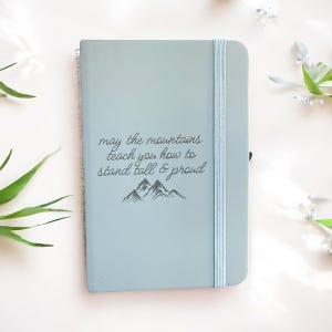 May include: A light blue notebook with a matching elastic band closure. The cover features the text "may the mountains teach you how to stand tall & proud" above a mountain graphic. The notebook is spiral bound.