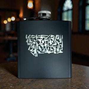 May include: A black stainless steel flask with a silver cap and a hinged closure. The front of the flask features a silver graphic of the state of Montana, composed of various gun silhouettes.