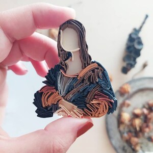 May include: A paper quilling art piece depicting the Mona Lisa. The artwork is made of layered strips of paper in shades of brown, orange, and blue. The piece is held in a hand with red nail polish.