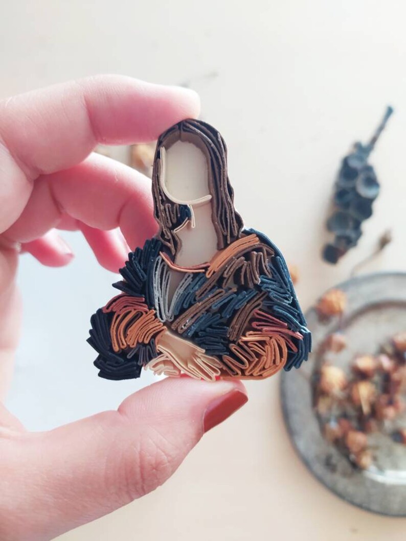 May include: A paper quilling art piece depicting the Mona Lisa. The artwork is made of layered strips of paper in shades of brown, orange, and blue. The piece is held in a hand with red nail polish.