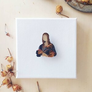 May include: A small canvas painting of the Mona Lisa, recreated using colorful yarn. The painting is on a white canvas and is surrounded by dried flowers.