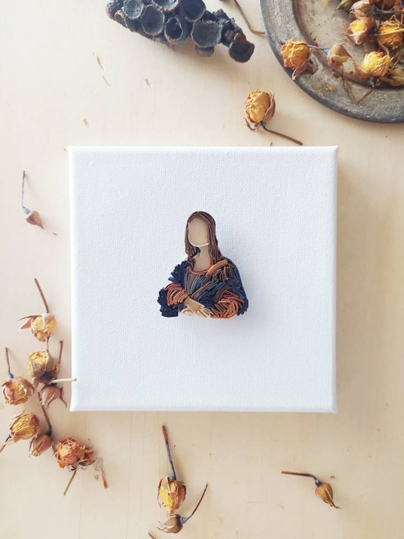 May include: A small canvas painting of the Mona Lisa, recreated using colorful yarn. The painting is on a white canvas and is surrounded by dried flowers.
