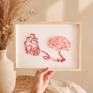 May include: A framed wall art piece featuring a red paper quilled anatomical heart and a human brain connected by a thin red line. The artwork is set against a white background within a light-colored wooden frame, showcasing intricate paper art.