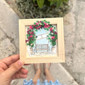 Puede incluir: Un marco cuadrado de madera clara que contiene una pieza de arte en filigrana de papel. La obra muestra una escena de jardín con un banco marrón bajo un arco floral de tiras de papel rosa y verde, flanqueado por dos macetas beige sobre fondo azul.