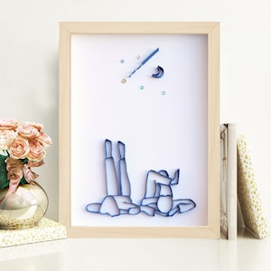 May include: A framed paper art piece depicting two figures lying on their backs, looking up at a crescent moon and stars. The figures are made of blue paper and are outlined in black. The background is white.