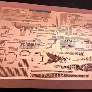 May include: A sheet of etched metal components for a model kit, featuring detailed outlines of various parts. The parts are arranged on a copper-colored sheet, with a brown cardboard backing. The design includes geometric shapes and mechanical elements.