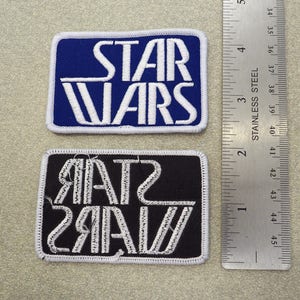 Puede incluir: Dos parches rectangulares bordados con el logotipo de Star Wars en hilo blanco. Un parche tiene fondo azul y el otro fondo negro. Una regla de acero inoxidable está colocada verticalmente al lado, mostrando una anchura de 7,6 cm.