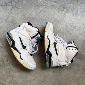 Op de afbeelding: Paar wit en zwarte high-top sneakers met een turquoise accent op de zolen. De sneakers hebben een zwarte Nike swoosh en het woord "FORCE" in turquoise. De schoenen staan op een grijze betonnen ondergrond.