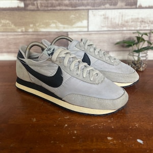 Pode incluir: Par de ténis Nike vintage cinzentos com logótipos pretos. Os sapatos têm atacadores e solas brancas, com uma faixa preta na parte inferior. Os ténis são exibidos numa superfície de madeira.
