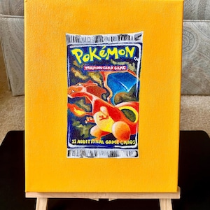 May include: A painting of a Pokémon Trading Card Game pack on a yellow canvas. The pack features a red and orange dragon-like creature. The canvas is displayed on a small wooden easel.