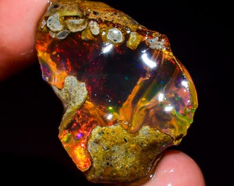 May include: A rough, natural opal gemstone with a fiery orange, red, and green play of color. The opal is set in a matrix of dark brown and gray rock.