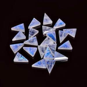 Può includere: Una collezione di gemme triangolari di pietra di luna. Ogni pietra ha una base bianca traslucida con riflessi blu interni. Le gemme sono disposte su uno sfondo scuro, evidenziando la loro bellezza naturale e i motivi unici.