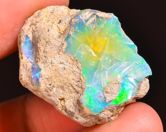 May include: A rough, uncut opal gemstone with vibrant colors of green, blue, and yellow. The opal is set within a beige, textured matrix. The gemstone is held in a person's hand.