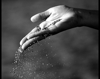 May include: A black and white photograph showing a hand with two thin metal rings on the fingers, with fine grains of sand pouring through the open palm and falling downward in a scattered stream against a dark, high-contrast background.