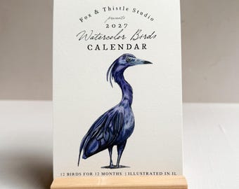 Könnte beinhalten: Ein Kalender für 2027 mit einer Aquarellillustration eines dunkelblauen Vogels. Der Text lautet Fox & Thistle Studio presents 2027 Watercolor Birds CALENDAR und 12 BIRDS FOR 12 MONTHS | ILLUSTRATED IN IL. Er steht in einem hellen Holzblock.
