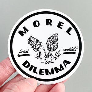 May include: Black and white sticker with a circle design. The text "MOREL DILEMMA" is written in a bold font around the circle. Inside the circle, there are two morel mushrooms with the text "fried" and "saut&eacute;d?" written above them.