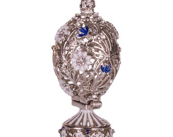 May include: Silver-toned, egg-shaped decorative box with intricate openwork and floral designs. The box features white and blue enamel accents, along with clear crystal embellishments. It has a hinged opening and sits on a pedestal base.