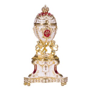 Puede incluir: Un huevo de Fabergé blanco y dorado con detalles de esmalte rojo y una corona en la parte superior. El huevo está sobre un soporte dorado con una base blanca y detalles intrincados. El huevo presenta un escudo rojo y dorado con la letra "M" en el centro.