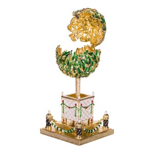 May include: A gold and green Christmas tree ornament with a white base and red accents. The ornament is in the shape of a tree with a round top and a square base. The tree is decorated with green leaves and red berries. The base is decorated with a white and red checkered pattern and a garland of green leaves and red berries.