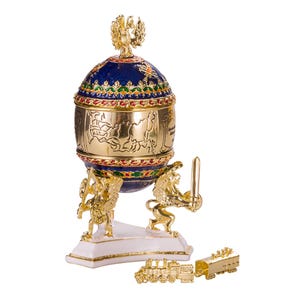 May include: A gold-colored decorative egg with a blue and gold top, supported by two golden lion figures. The egg features intricate detailing and a small golden train. The egg is a decorative item.