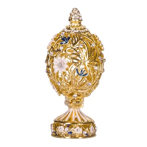Pode incluir: Um ovo decorativo dourado com um design filigranado e detalhes em esmalte branco e azul. O ovo é adornado com strass e fica em um suporte dourado.