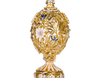 Op de afbeelding: Een decoratief gouden ei met een filigraanontwerp en witte en blauwe emaille accenten. Het ei is versierd met strass en staat op een gouden standaard.