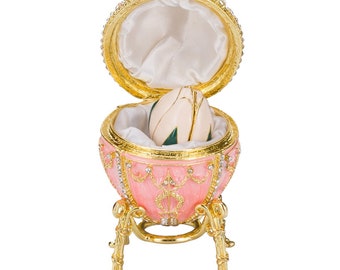 May include: A pink, egg-shaped decorative box with gold accents and a hinged lid. The box is adorned with rhinestones and rests on a gold stand. Inside, a white and green egg-shaped object sits on a white satin lining.
