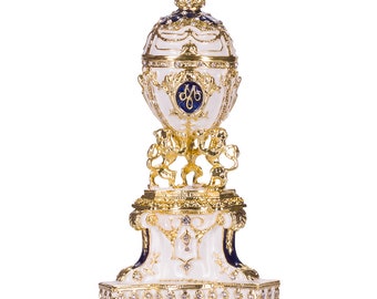 Puede incluir: Un huevo de Fabergé blanco y dorado con una parte superior de esmalte azul y una corona. El huevo está sobre un soporte dorado con una base blanca y detalles azules. El huevo está decorado con detalles intrincados y joyas.