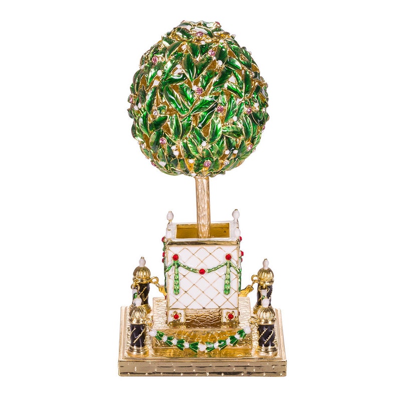 May include: A gold-tone decorative egg with a green and white enamel tree design. The tree has a round top and is set on a gold-tone base with white and red accents.