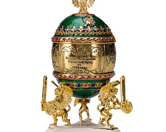 May include: A decorative egg-shaped ornament with a green and gold exterior, supported by three golden lion figures. The egg features intricate detailing, including text and colorful enamel accents. A golden double-headed eagle tops the ornament.