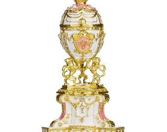 Op de afbeelding: Een witte en gouden Fabergé-eireplik met een roze en gouden kroon bovenop. Het ei is versierd met gouden randen en een roze en gouden wapenschild. Het ei staat op een gouden standaard met een witte en gouden basis.