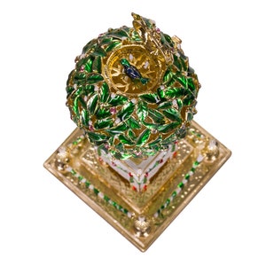 May include: A gold-colored decorative object with a green and white top, resembling a tree with a bird on top. The base is a square with a gold-colored border and a white center.