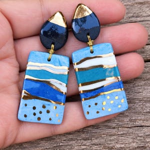Può includere: Un paio di orecchini pendenti con una parte superiore a goccia blu scuro con punta dorata. La parte rettangolare inferiore presenta strisce orizzontali azzurre, turchesi e bianche, arricchite da linee dorate e piccoli punti dorati.