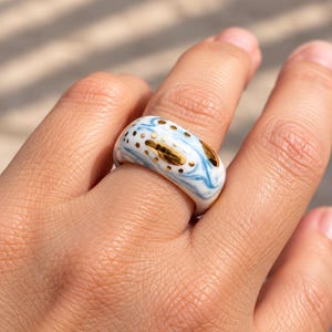 May include: A wide ceramic ring featuring a white base with swirling light blue marbled patterns. The surface is decorated with metallic gold accents, including small dots and elongated shapes, creating a distinct contrast against the blue and white.