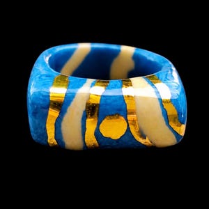 May include: A chunky ring featuring a polished blue base with irregular cream-coloured bands and thin, reflective gold-toned metallic stripes integrated into the surface. The ring has a rounded square shape with a smooth, glossy finish throughout.