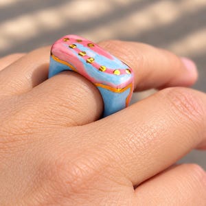 May include: A chunky rectangular ring featuring a smooth, glossy finish with a marbled pattern of pastel pink and light blue. The surface is accented with a thin, wavy gold-colored line and a series of small, circular gold-colored dots.