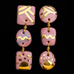 May include: A pair of dangling earrings featuring three stacked ceramic pieces in shades of mauve and pink. Each piece is decorated with gold-colored metallic accents, including lines, waves, and dots, and connected by thin gold-colored wire links.