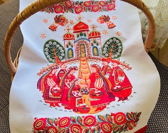 May include: White linen kitchen towel with a red embroidered border and a colorful folk art design featuring a church, trees, and people in traditional clothing. The design is surrounded by a border of red and green flowers.
