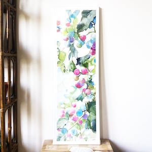 May include: Watercolor painting of a botanical scene with pink, blue, and purple berries and green leaves. The artwork is framed in a white rectangular frame and rests on a wooden surface. The painting has a soft, airy feel.