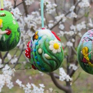 Könnte beinhalten: Drei Filz-Ostereier mit Frühlingsmotiven. Jedes Ei zeigt ein Mädchen, einen Schmetterling und Blumen. Die Eier hängen an einem Ast.
