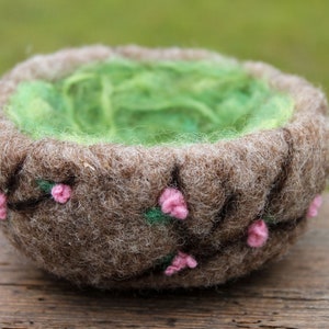 Könnte beinhalten: Eine braune Filzschale mit grüner Filzfütterung und rosa Filzblumen. Die Schale ist handgefertigt und hat einen rustikalen Look.
