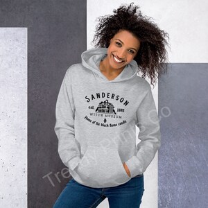 Op de afbeelding: Grijs hoodie met een zwarte grafische print van een huis met de tekst "Sanderson est. 1693 Witch Museum Home of the black flame candle"