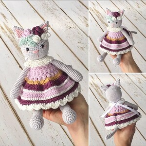 Könnte beinhalten: Handgefertigte Häkelkatzenpuppe mit grauem Körper und einem bunten Kleid. Das Kleid hat Streifen in Rosa, Lila und Gelb mit weißer Spitzenborte. Die Katze hat ein Blumenstirnband und einen freundlichen Gesichtsausdruck.