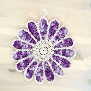 Könnte beinhalten: Ein runder, blumenförmiger Topflappen aus Häkelarbeit mit einer cremefarbenen Mitte, umgeben von zwölf Blütenblättern in verschiedenen Violetttönen, von hellem Lavendel bis zu dunklem Lila, eingefasst mit einem cremefarbenen Garnrand und einer Schlaufe.