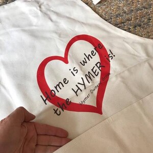 May include: White canvas apron with a red heart and the text "Home is where the Hymer is!" printed on it. Hymer Owners Group is printed in smaller text below the heart.
