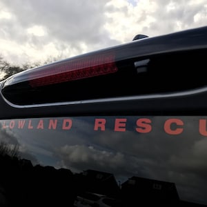 Puede incluir: Un vehículo negro con una luz trasera roja y una pegatina roja "LOWLAND RESCUE" en la ventana trasera.