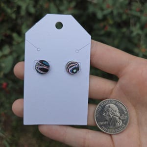 May include: Two round stud earrings with a swirling pattern of black, blue, and iridescent colors are displayed on a white earring card. A US quarter coin is next to the earrings for size comparison. The earrings have a unique, eye-catching design.