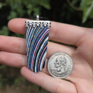 May include: A rectangular pendant with a silver-colored top and a colorful striped design in shades of blue, black, pink, and white. The pendant is held next to a U.S. quarter coin.