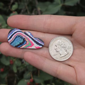 May include: A cloud-shaped brooch with a colorful, layered design. The brooch features shades of blue, pink, green, and black. A US quarter coin is next to the brooch for size comparison. The brooch is held in a hand.