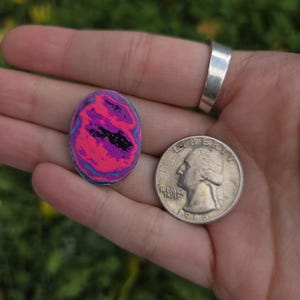 May include: An oval-shaped, colorful art piece with a swirling pattern of pink, purple, black, and blue. A silver quarter coin is next to the art piece for size comparison. The art piece is held in a hand.